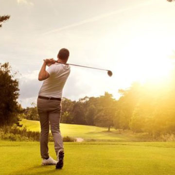 A man in a white shirt and gray pants swings a golf club on a green golf course at sunset, with trees and sunlight in the background.