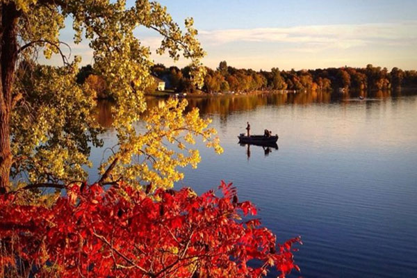 A small boat with two people floats on a calm lake, surrounded by trees with colorful autumn leaves in shades of red, yellow, and green, under a clear sky.