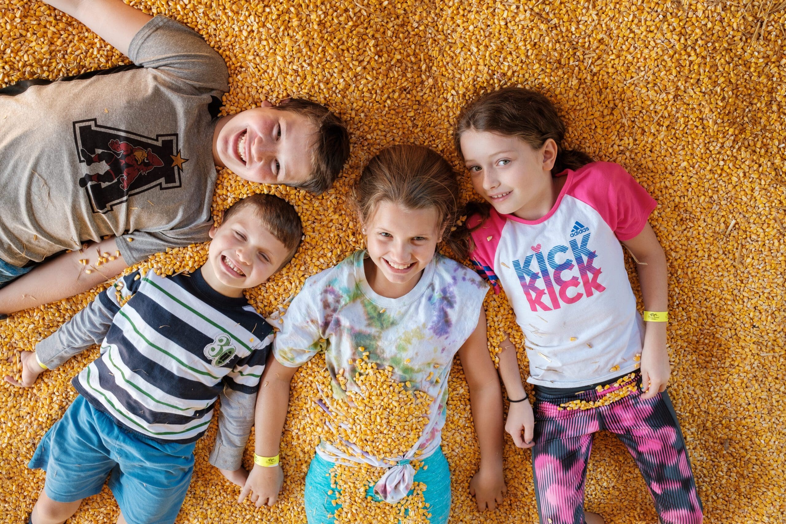 Four children lie side by side, smiling up at the camera on a bed of yellow corn kernels at Country Blossom Farm. Dressed in colorful, casual clothes, they radiate joy and playful energy in this cheerful moment.