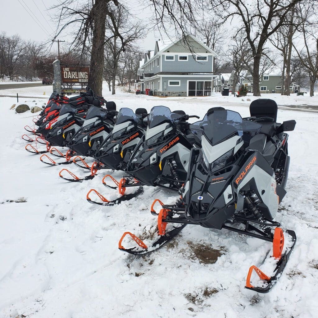A row of Polaris snowmobiles with orange skis is parked on snow in front of a gray building with the sign "Lake Darling" at Lake Darling Resort. Bare trees and scenic winter views complete the backdrop.