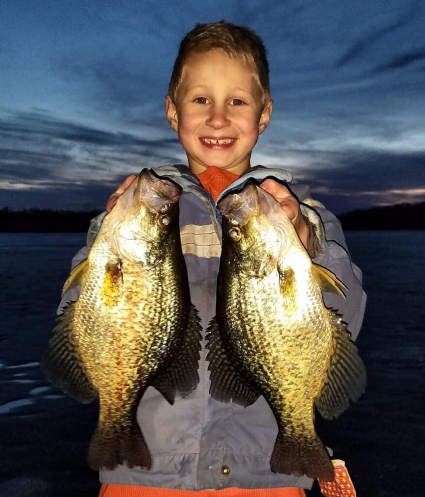 A smiling young boy stands outdoors at dusk, holding up two large, shiny fish caught during an aggressive bite, one in each hand. The sky behind him is darkening with streaks of clouds.
