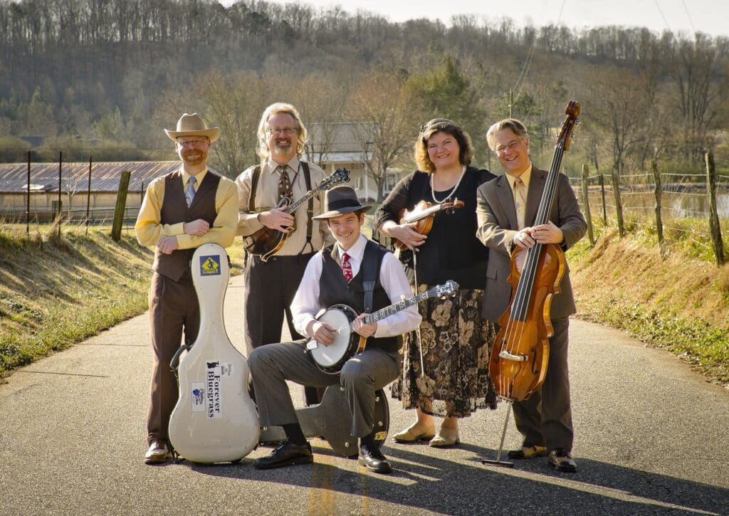 Five musicians pose on a rural road with their instruments and vintage-style outfits, smiling amid trees and a barn—capturing the charm of early spring events in Alexandria, MN.