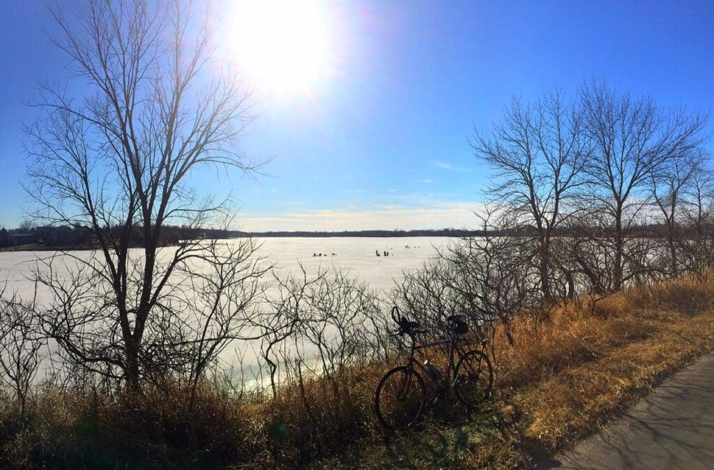 A bicycle rests on a sunlit path in Alexandria beside leafless trees, overlooking a frozen lake where people ice fish beneath a clear blue sky as spring arrives with clear trails.