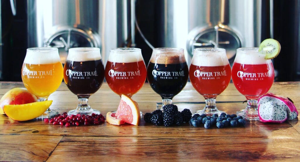 Six glasses of craft beer from Copper Trail Brewing Co, each a different color, are lined up on a wooden table with fresh mango, pomegranate, grapefruit, blackberry, blueberry, and dragon fruit in front. Brewing tanks appear in the background.