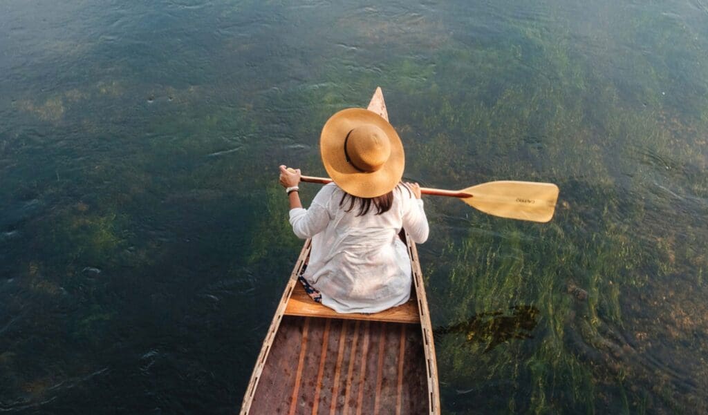 A person wearing a wide-brimmed hat paddles a wooden canoe on clear, calm water in the Lakes Area—a must-do for summer fun, viewed from above.