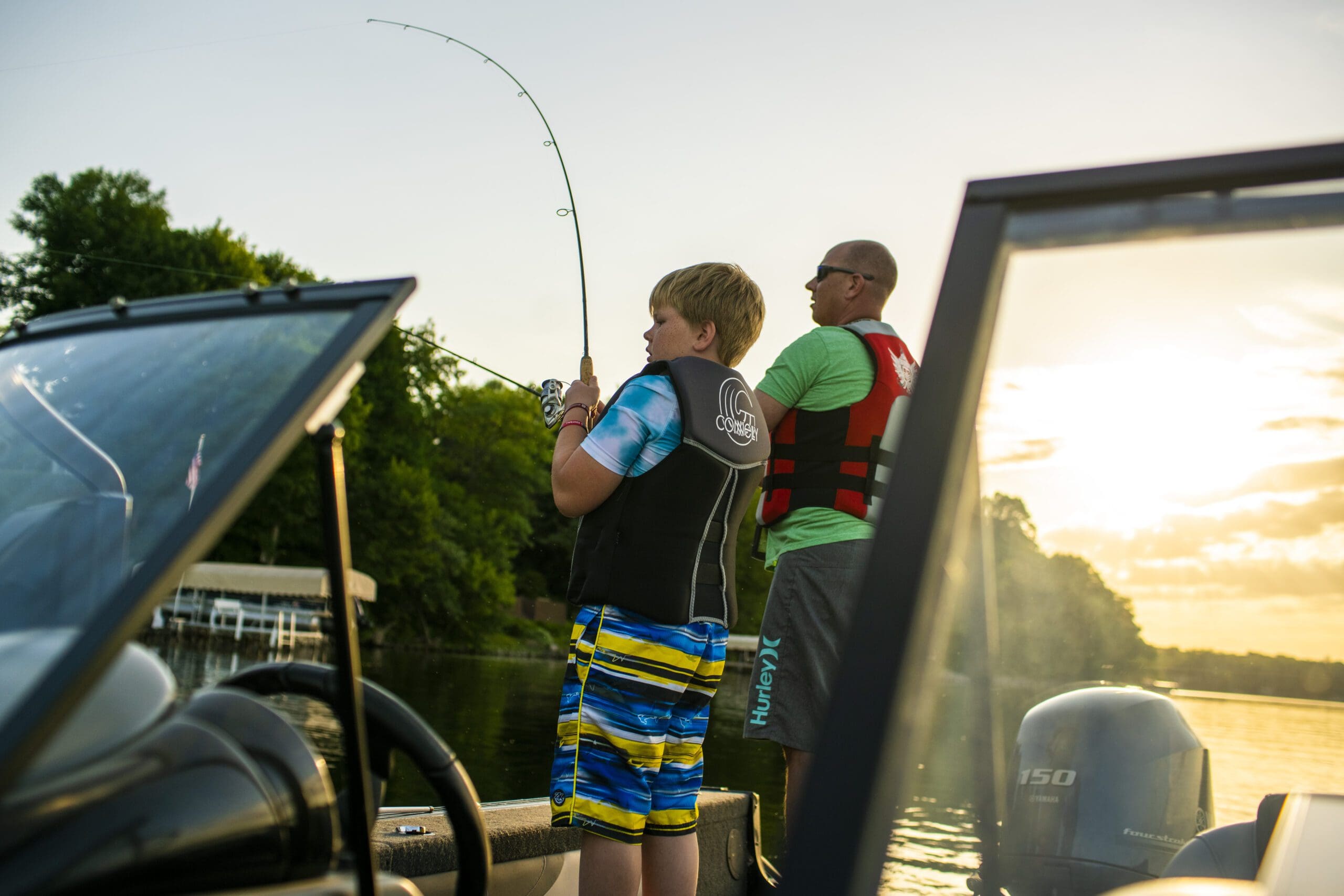 A boy and an adult wearing life jackets fish from a boat on a calm lake at sunset during their Alexandria getaway, with trees and a dock visible in the background.
