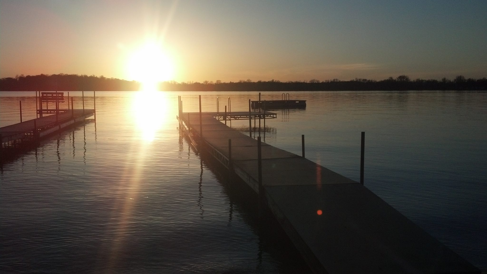 A bright sun sets over a calm lake in the Lakes Area, casting reflections on the water. Two empty docks extend into the lake, while trees line the distant shore under a clear sky—perfect for tourism during National Tourism Week.