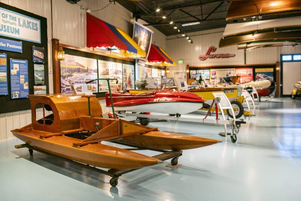 A museum exhibit in Alexandria Getaway features vintage wooden and fiberglass boats, colorful awnings, informational displays, and bright lighting in a spacious indoor setting.