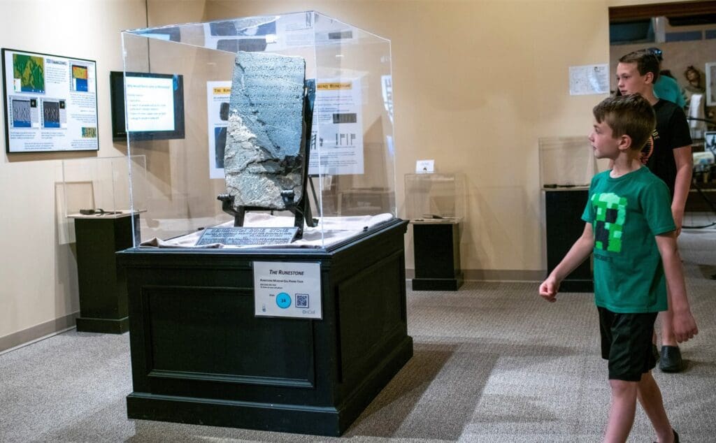 Two children view a large stone artifact displayed in a glass case at a museum exhibit, making their Alexandria getaway both educational and inspiring. Informational panels about the artifact are mounted on the walls behind them.