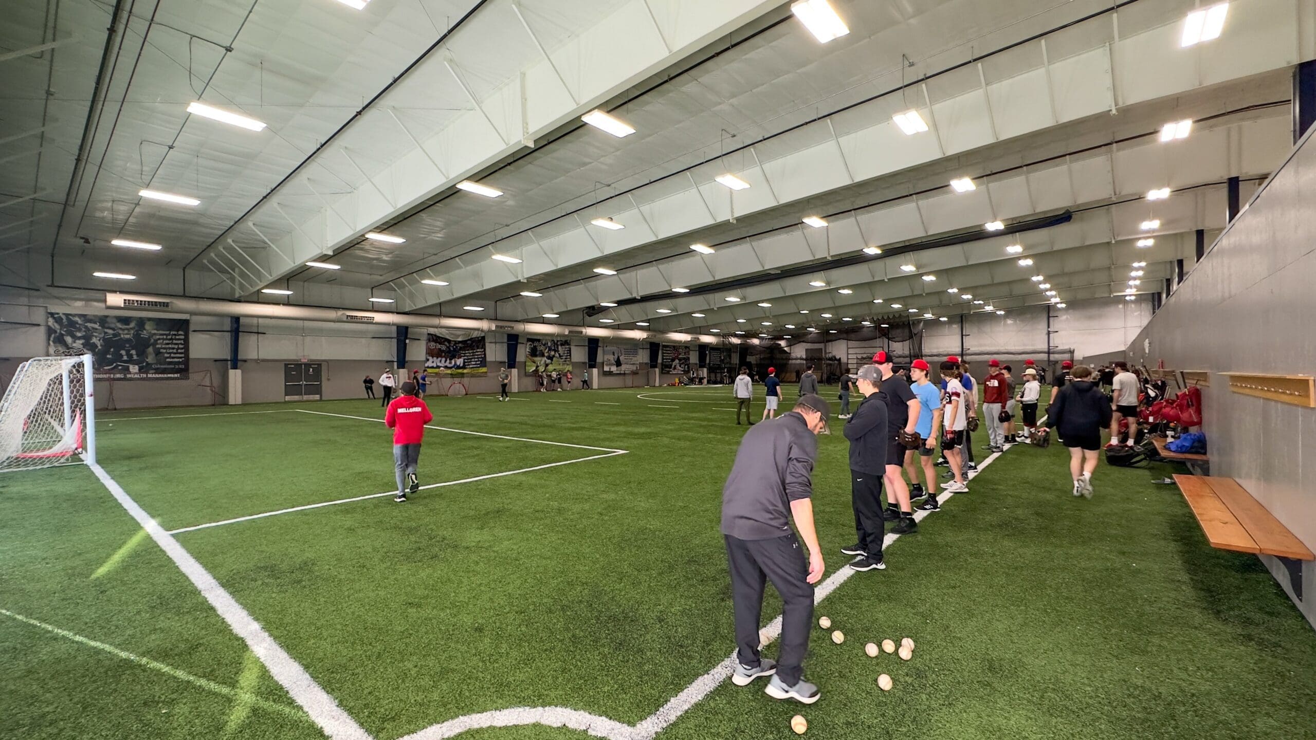 A group of people, mostly in athletic wear, stand in lines on an indoor turf field at NorthStar Sports Complex. Baseballs are scattered on the ground, and a netted goal is visible on the left. The large facility is well-lit with overhead lights.