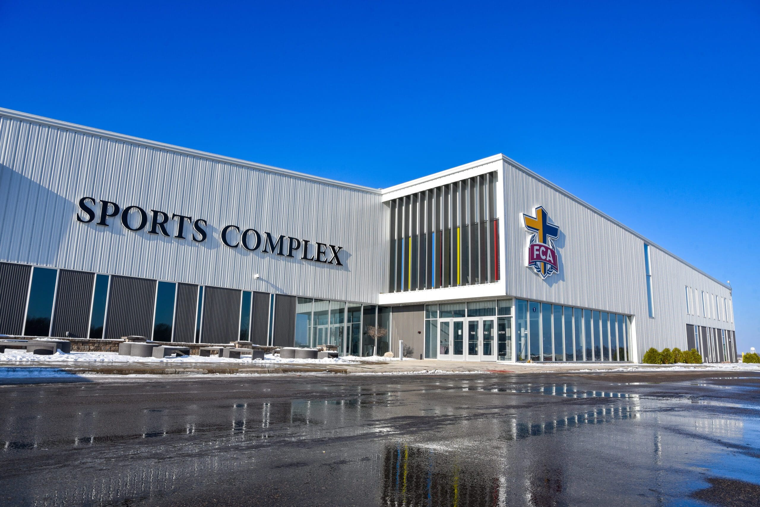A modern NorthStar Sports Complex building with large vertical windows and "SPORTS COMPLEX" and the "FCA" logo on the exterior. Snow and wet pavement are visible in front under a clear blue sky.