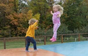 Two young girls play on an outdoor inflatable pad in the crisp air, one standing in a yellow top and jeans as the other leaps midair. Colorful fall trees set a lively scene—perfect for families looking to explore Alexandria MN.
