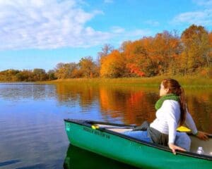 A woman sits relaxed in a green canoe on a calm lake, surrounded by colorful autumn trees reflected in the water under a partly cloudy blue sky—an inviting scene to explore Alexandria MN.