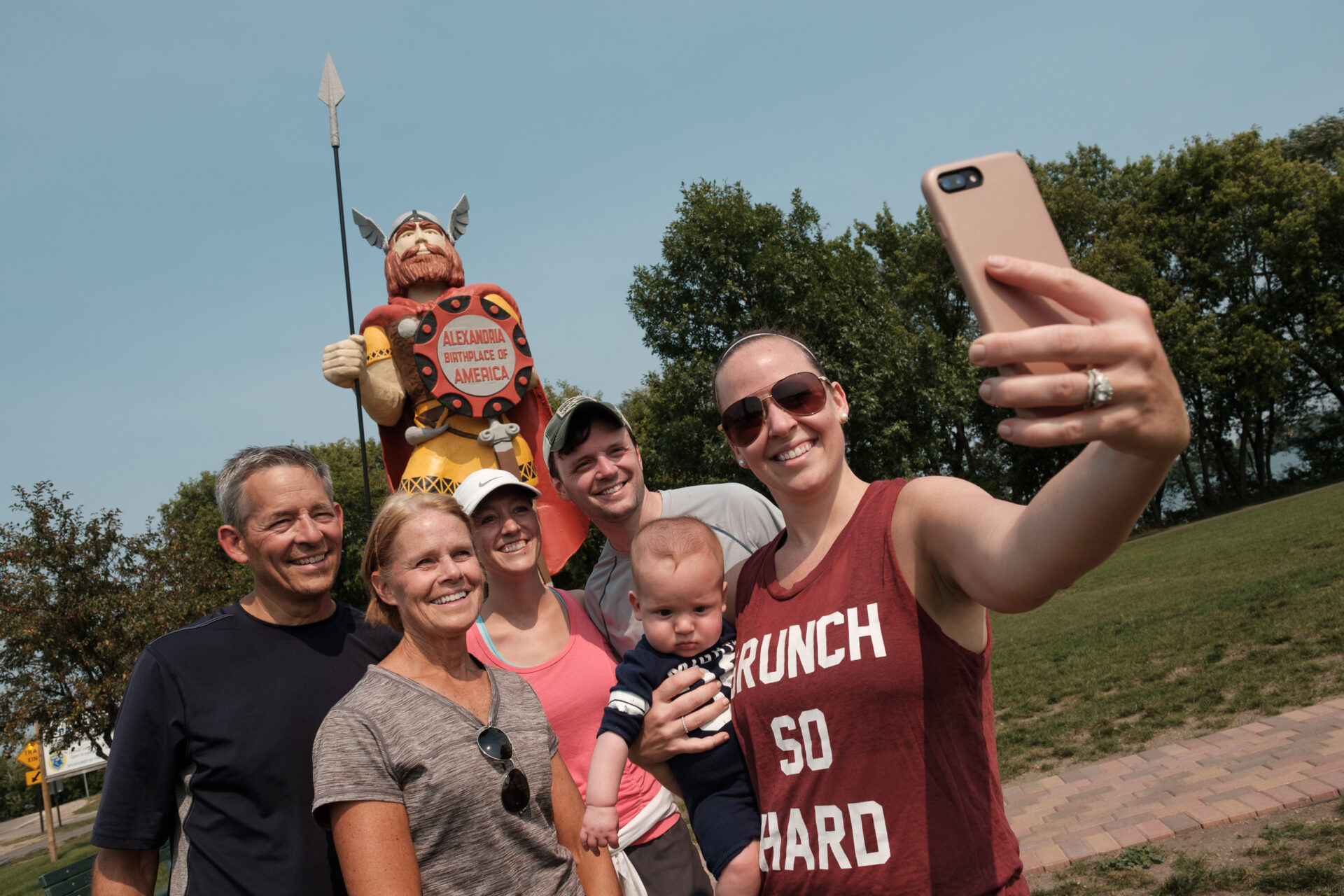 A group of six people, including a baby, smile and pose for a selfie in front of a Viking statue holding a spear and shield. Trees and a clear sky are in the background as they explore Alexandria MN.