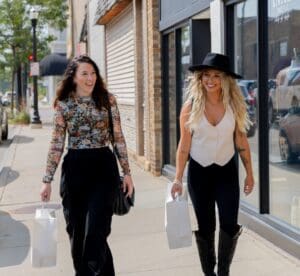 Two women smiling and walking on a city sidewalk, each carrying white shopping bags. One wears a floral top and black skirt, the other a white vest, black pants, and a black hat—ready to explore Alexandria MN among shops and tree-lined streets.