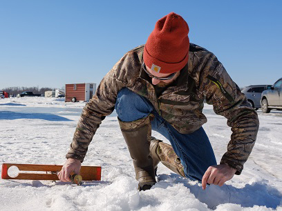A person in a camouflage jacket and red beanie uses a measuring tool on snow-covered ground, with vehicles and trailers in the background under a clear blue sky—perfect scenery to explore Alexandria MN.