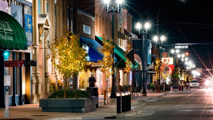 A well-lit downtown street at night in Alexandria, MN features trees decorated with lights, colorful storefront awnings, and empty sidewalks—creating a peaceful and inviting atmosphere to explore Alexandria, MN.
