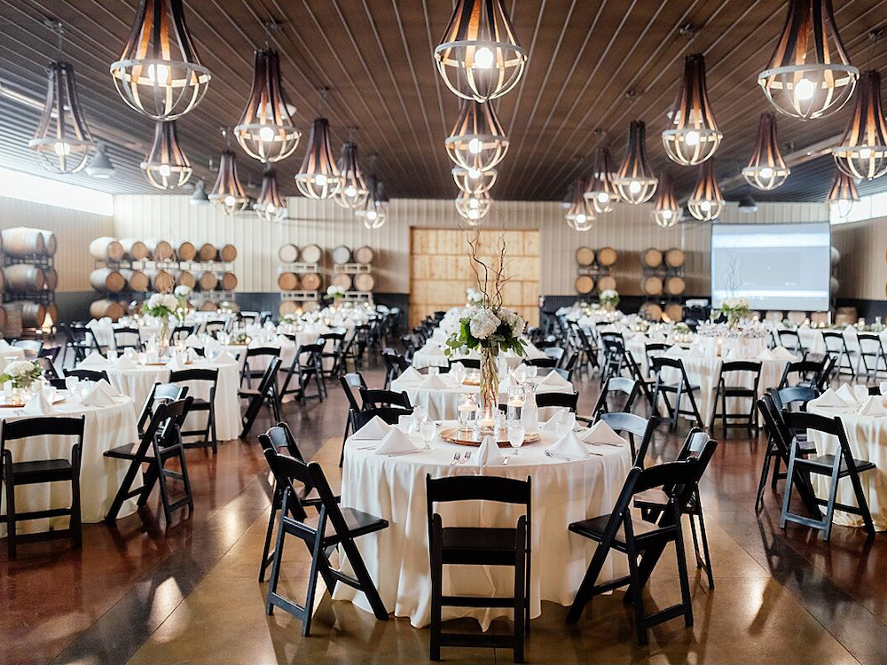 A spacious banquet hall with round tables covered in white tablecloths, black chairs, elegant floral centerpieces, wine barrels along the walls, and modern pendant lights hanging from a wooden ceiling.