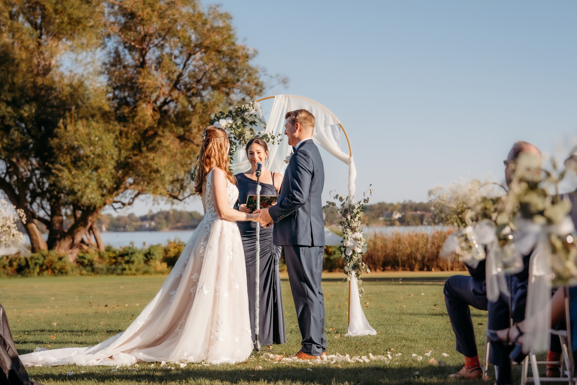 A bride and groom stand facing each other, holding hands during an outdoor wedding ceremony by a lake, with an officiant between them and trees in the background. Guests are seated on either side.