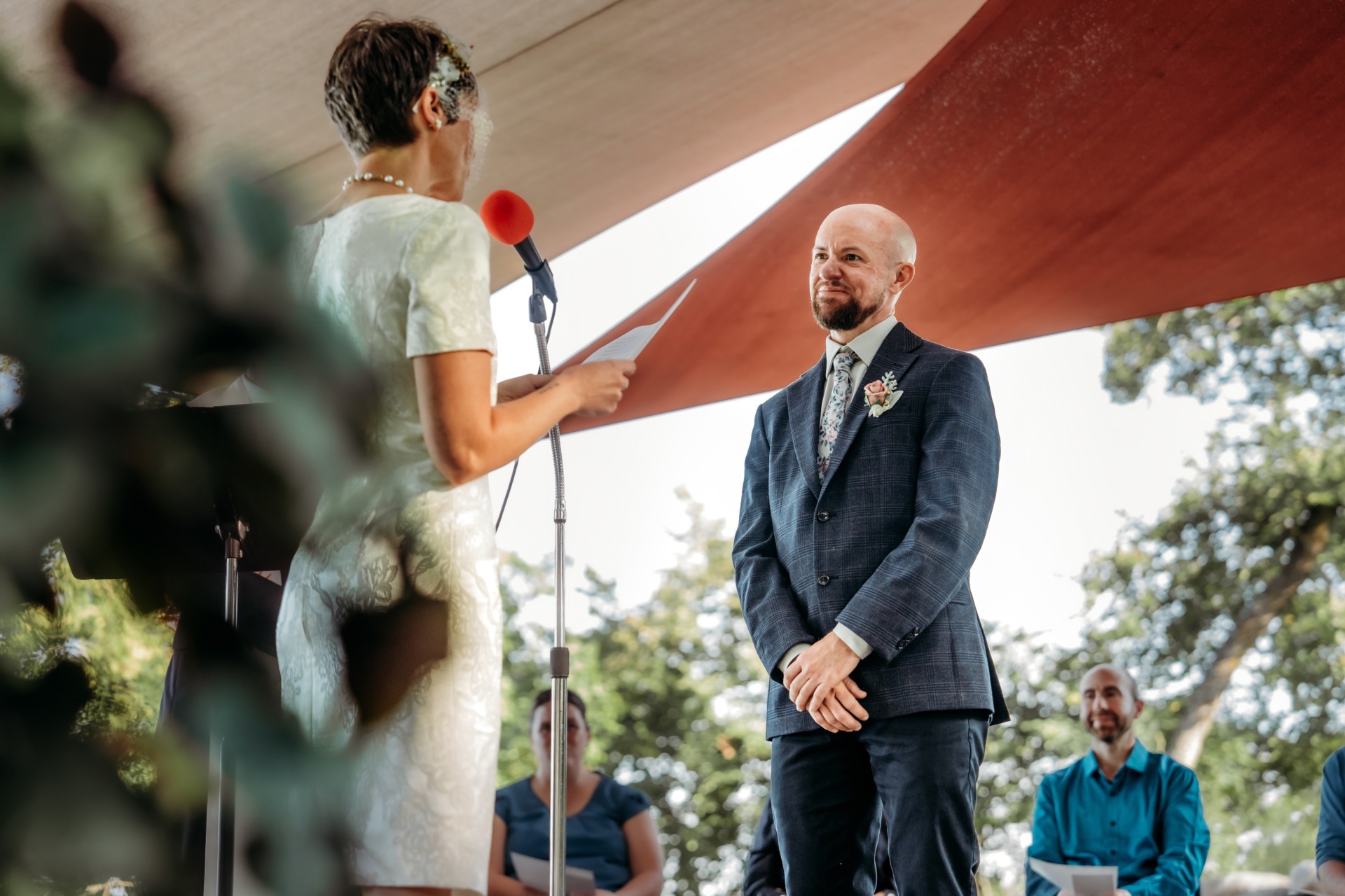 A woman in a light dress reads from a paper with a red microphone, while a man in a dark suit and floral boutonniere smiles at her during an outdoor ceremony under a canopy. Two seated guests watch in the background.
