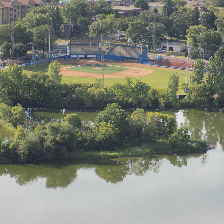A baseball field, often hosting sporting events, sits with empty stands surrounded by trees beyond a body of water edged in green foliage. In the distance, residential buildings and streets complete the background.
