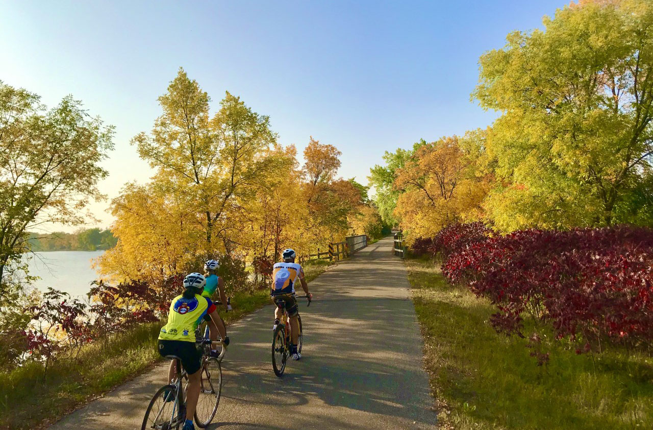 Three cyclists ride on a paved trail surrounded by colorful autumn trees with yellow, orange, and red leaves under a clear blue sky. A body of water is visible to the left of the path.