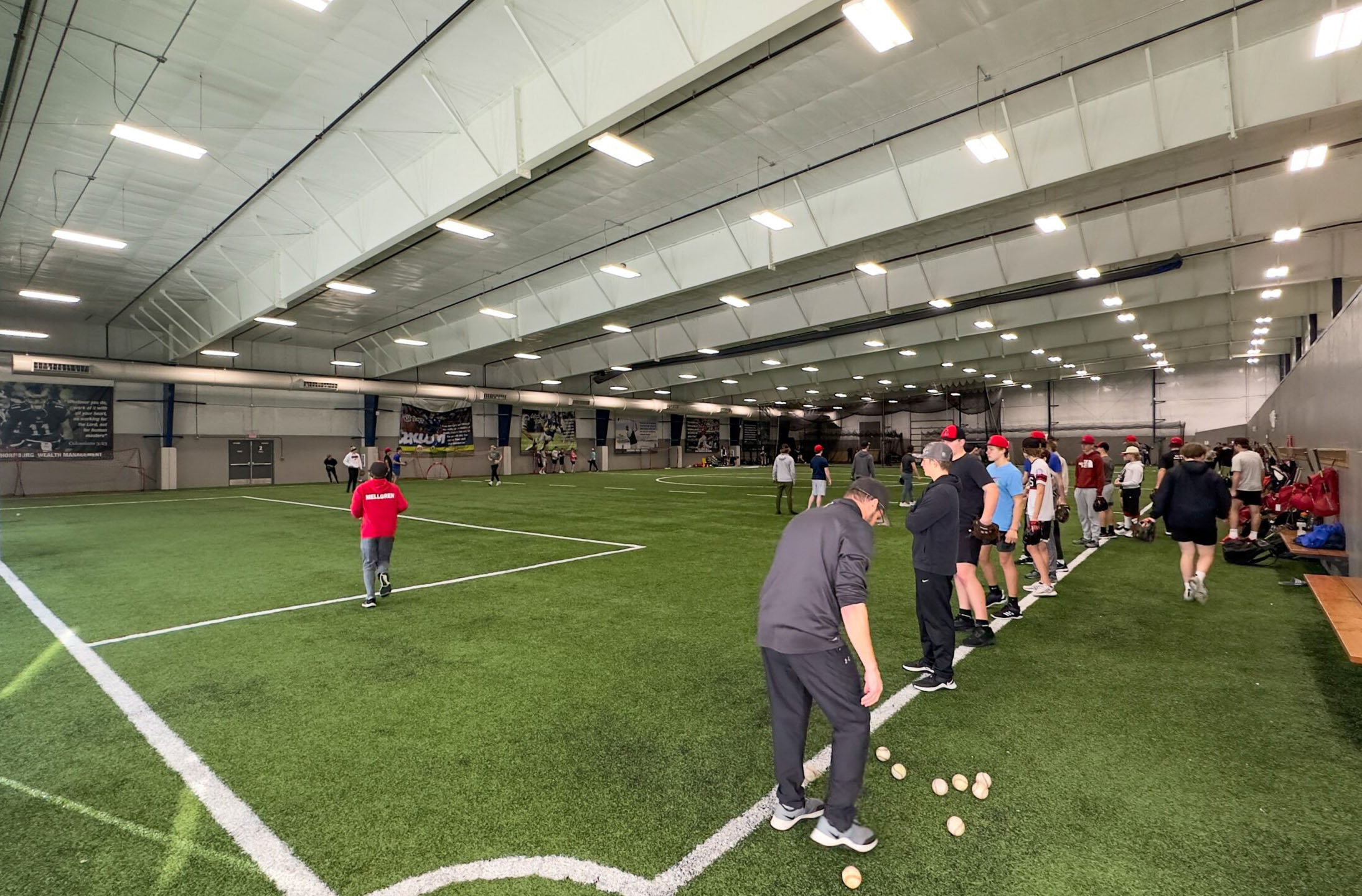 A group of kids and coaches in baseball gear gather along the sideline of an indoor turf field, with practice equipment and baseballs scattered nearby. The spacious, well-lit facility is perfect for training sessions or Special Events.