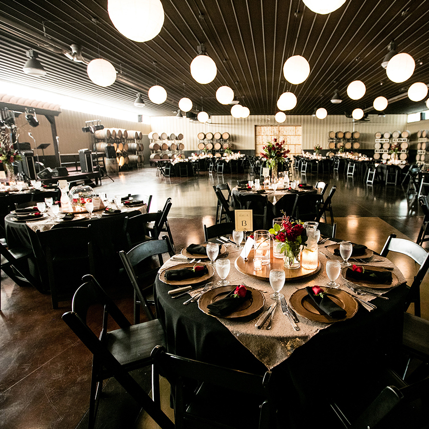 Elegant banquet hall with round tables set for conventions or meetings, featuring black tablecloths, floral centerpieces, wine barrels along the walls, and glowing spherical pendant lights hanging from the ceiling.