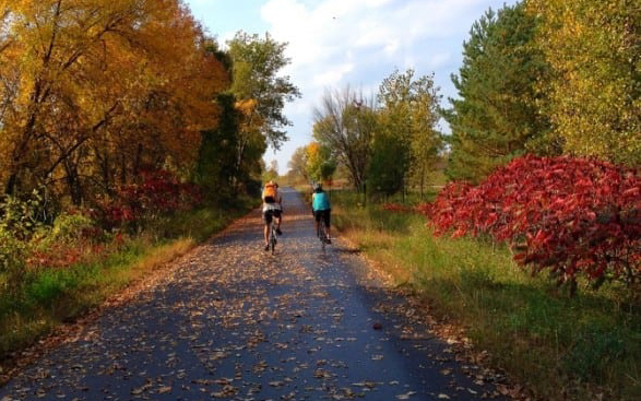 Two people ride bicycles down a paved trail surrounded by colorful autumn trees and scattered fallen leaves under a partly cloudy sky.