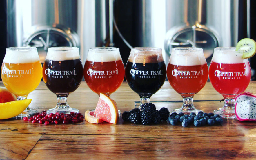 Six glasses of beer from Copper Trail Brewing Co., each paired with a different fruit: mango, pomegranate, grapefruit, blackberry, blueberry, and dragon fruit, displayed on a wooden table with brewery tanks in the background.