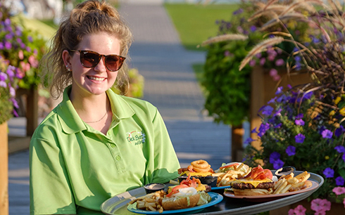 A smiling woman wearing sunglasses and a green polo shirt holds a tray with burgers, fries, onion rings, and other food, standing outdoors near colorful flowers.