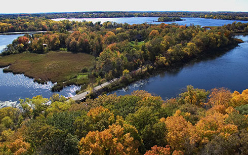 A scenic aerial view of a narrow road bordered by trees with autumn foliage, surrounded by blue lakes and wooded islands under a clear sky.