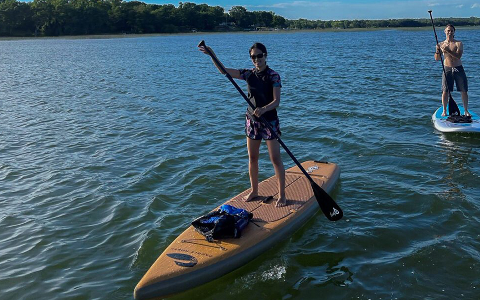 Two people are stand-up paddleboarding on a lake under a clear blue sky. The woman in the foreground wears sunglasses and a life jacket, while a man in the background paddles on a separate board. Trees line the shore.