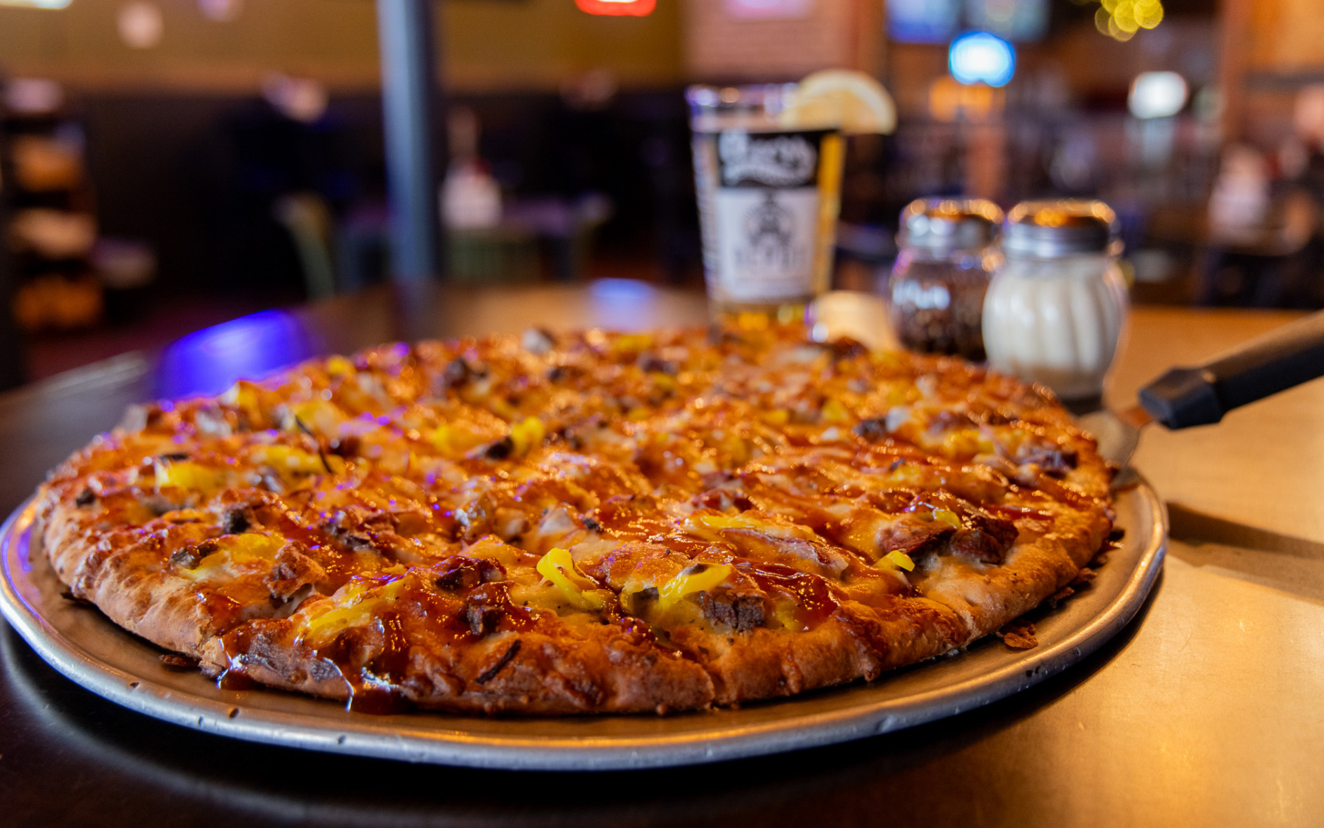 A freshly baked pizza topped with cheese, beef, onions, and pepper rings sits on a metal tray—a cozy winter guide essential—alongside a glass of beer, salt and pepper shakers, and a blurred restaurant background.