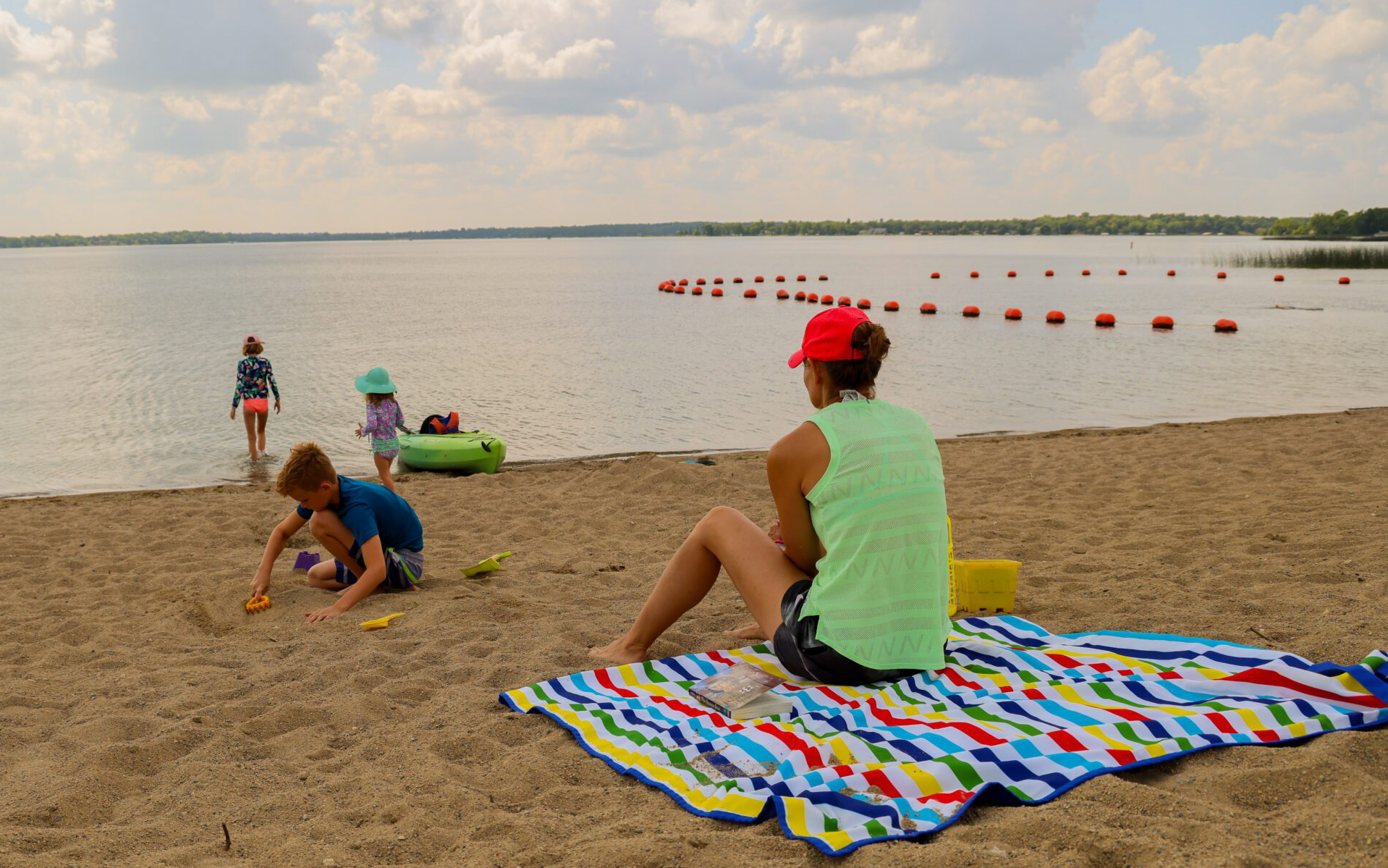 A woman sits on a colorful striped blanket on a sandy beach, watching children play nearby as outlined in the Summer Guide. Cloudy skies loom above, orange buoys dot the lake, and laughter fills the air.