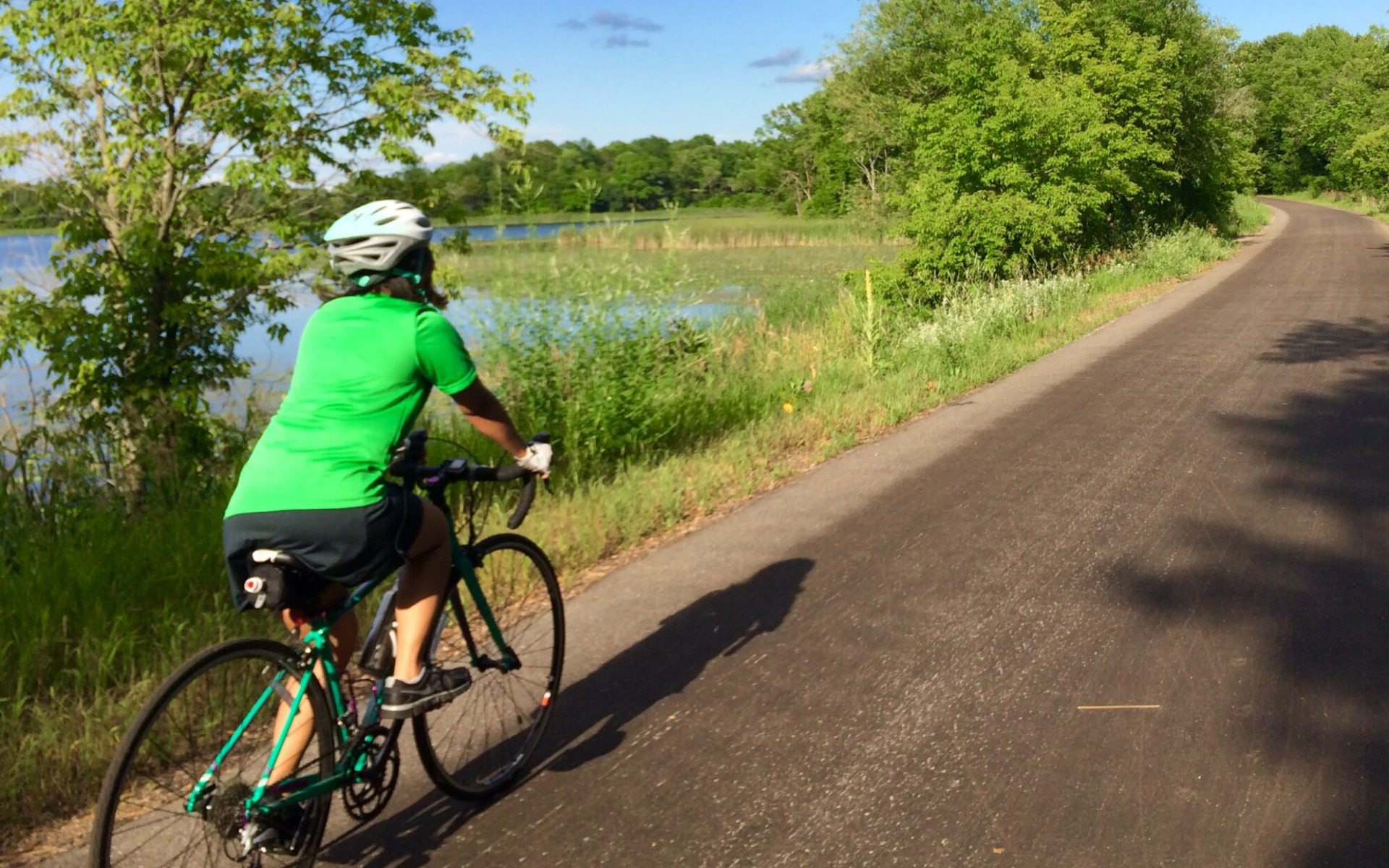 A person in a green shirt and white helmet rides a bicycle on a paved path next to a lake, surrounded by green trees and grass under a blue sky—a perfect scene for any Summer Guide.