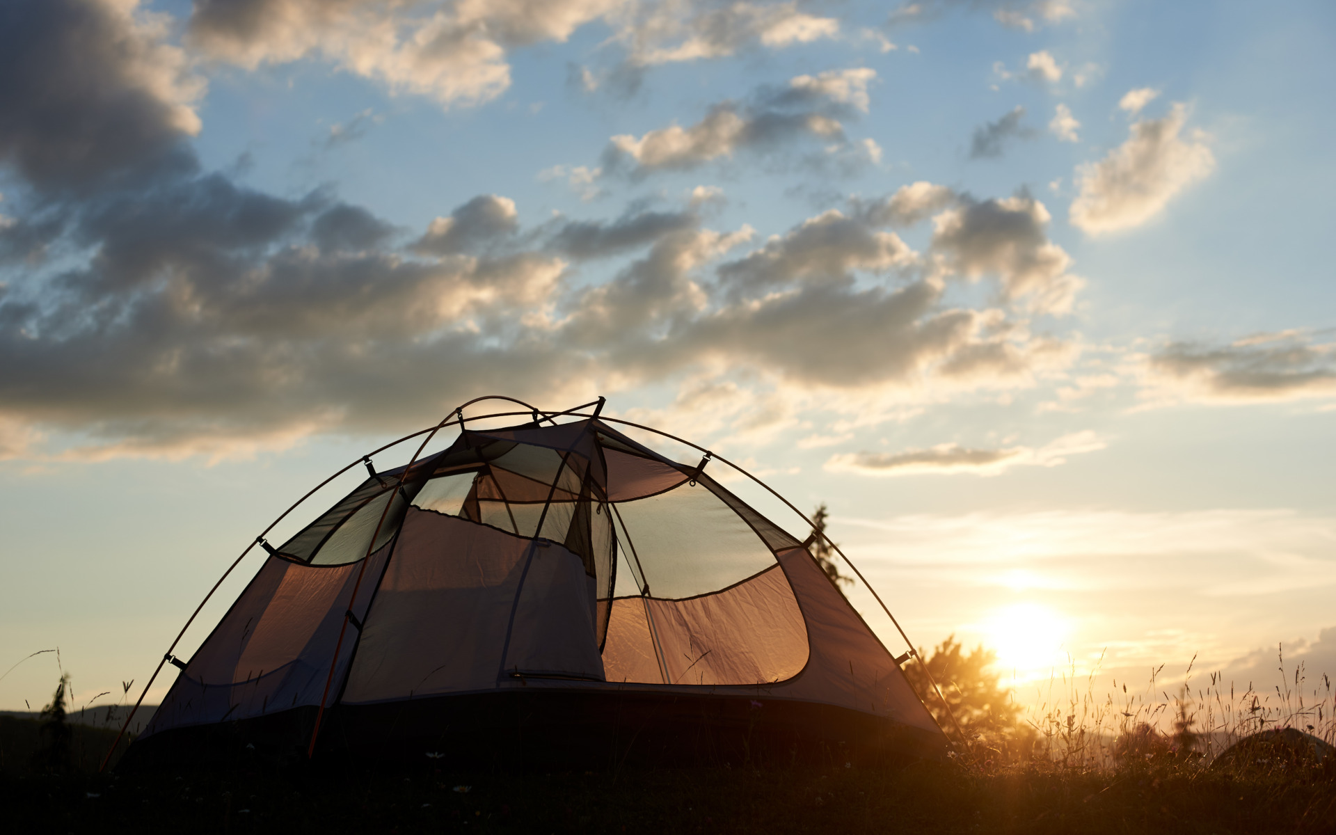 A dome-shaped tent set up on grass is silhouetted against a cloudy sky at sunset, with soft sunlight illuminating the scene—a perfect moment for any Summer Guide.