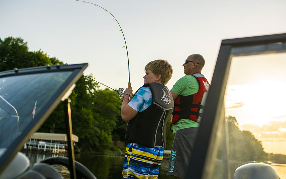A child wearing a life jacket and colorful shorts fishes with an adult in a red life vest on a boat at sunset, surrounded by calm water and trees—a perfect moment from any Summer Guide.