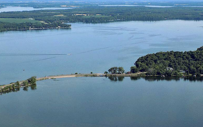 Aerial view of a lake with a thin, tree-lined peninsula extending into the water. A boat leaves a wake on the lake, surrounded by green fields and forests—an inviting scene for any Summer Guide adventure.