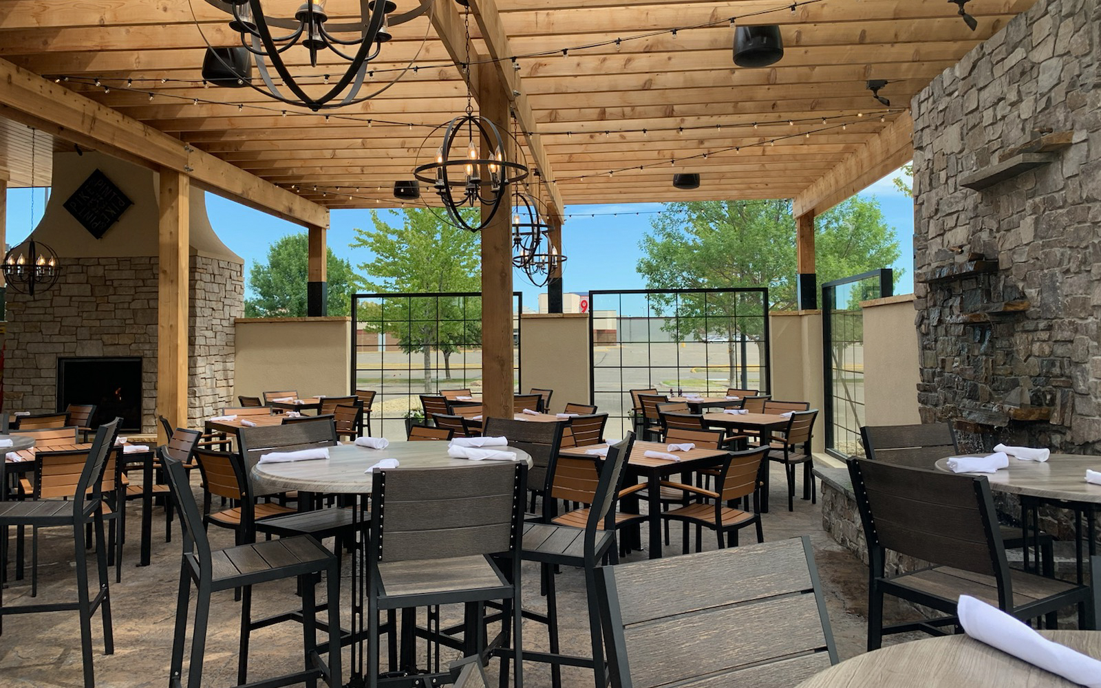 Covered outdoor restaurant patio with wooden beams, stone walls, and string lights sets the scene for a perfect evening. Multiple tables and chairs are neatly arranged—an ideal spot featured in any Summer Guide—with trees and a parking lot visible through clear panels.
