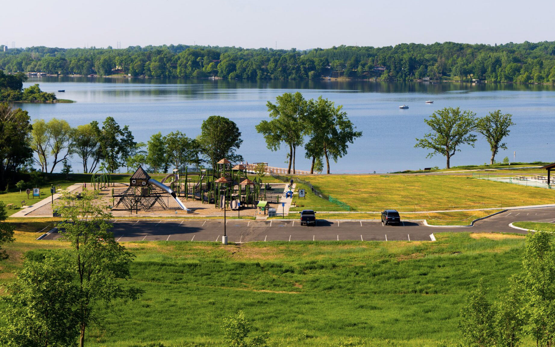 A scenic lakeside park featured in our Summer Guide, with a playground, open grassy areas, a parking lot with a few cars, and a dock extending into a calm lake surrounded by green trees on a sunny day.