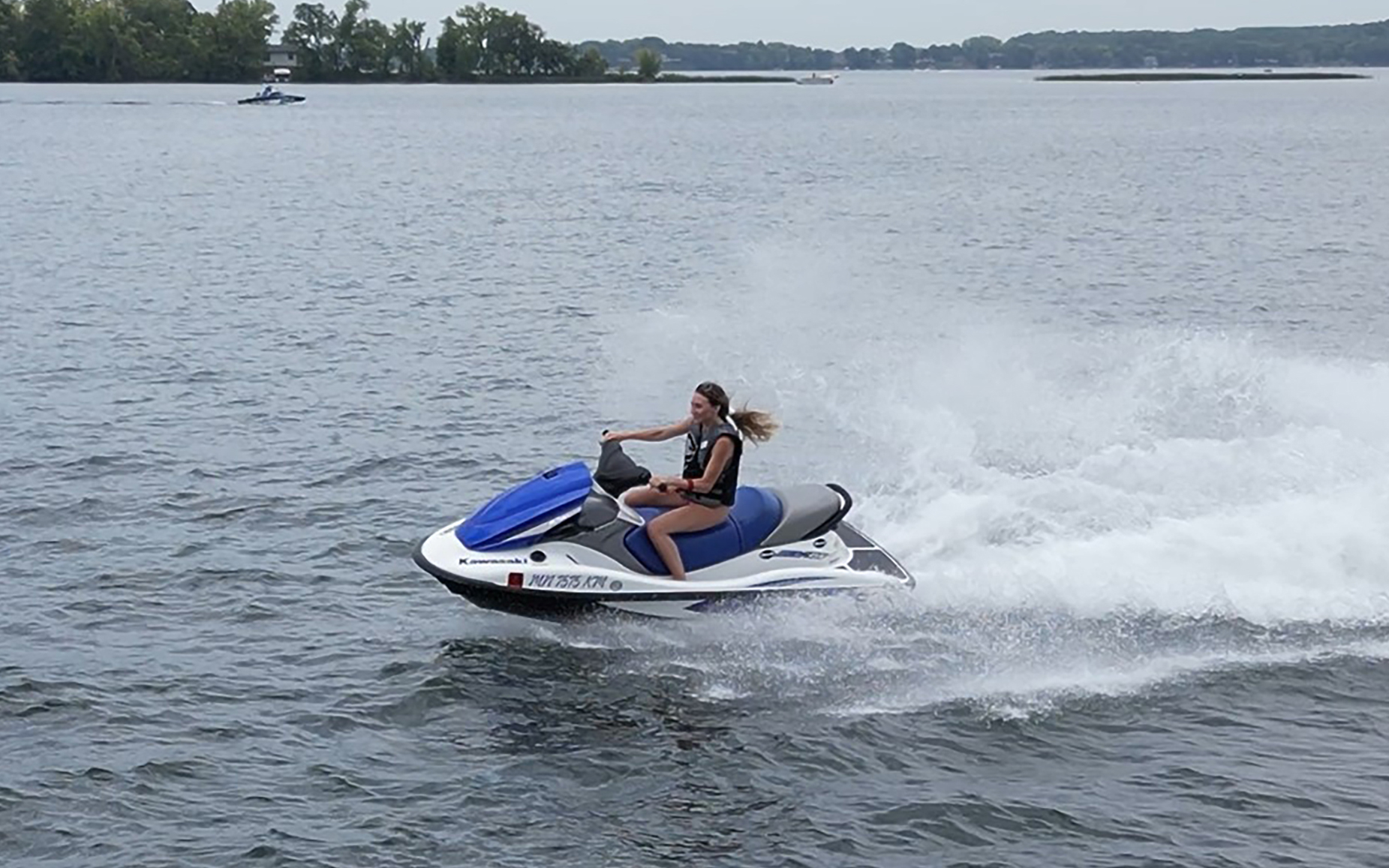 A person rides a blue and white jet ski at high speed across a large, calm lake in this Summer Guide moment, creating a spray of water behind them; trees and distant land are visible in the background.