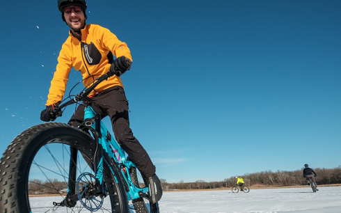 A person in a yellow jacket rides a fat-tire bike on a snowy or icy surface under a clear blue sky, Winter Guide style, with two other cyclists pedaling in the background.