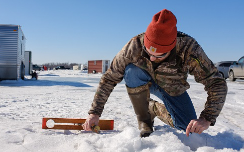 A Winter Guide in a camouflage jacket and orange beanie kneels on snowy ground, using a tool to measure ice thickness near ice fishing shelters and parked vehicles under a clear blue sky.