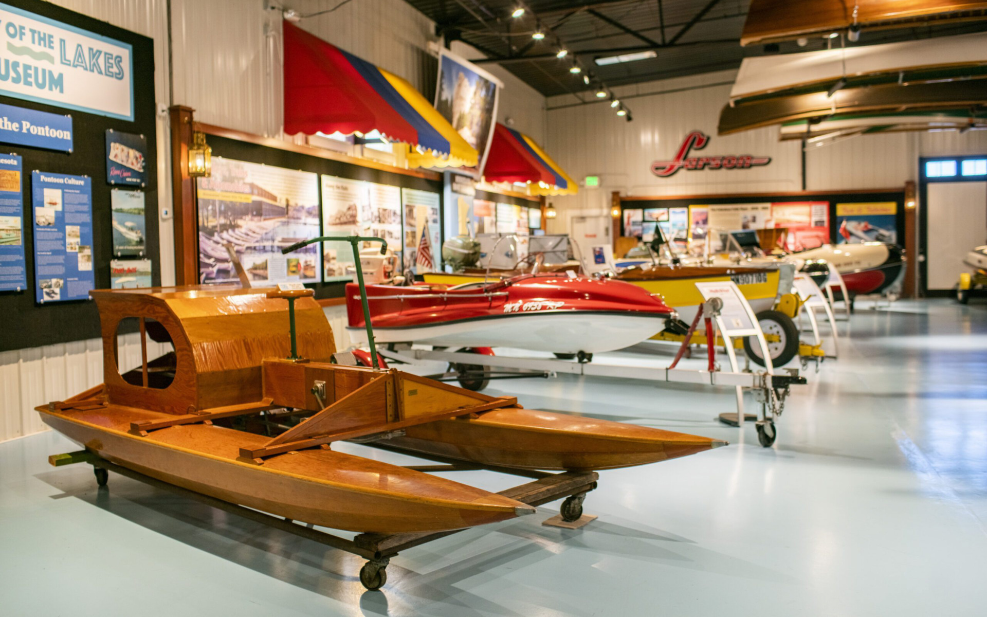 A Winter Guide museum exhibit displays vintage wooden boats and colorful speedboats, with informational panels in the background. The boats are arranged indoors on a shiny blue floor under bright lighting.