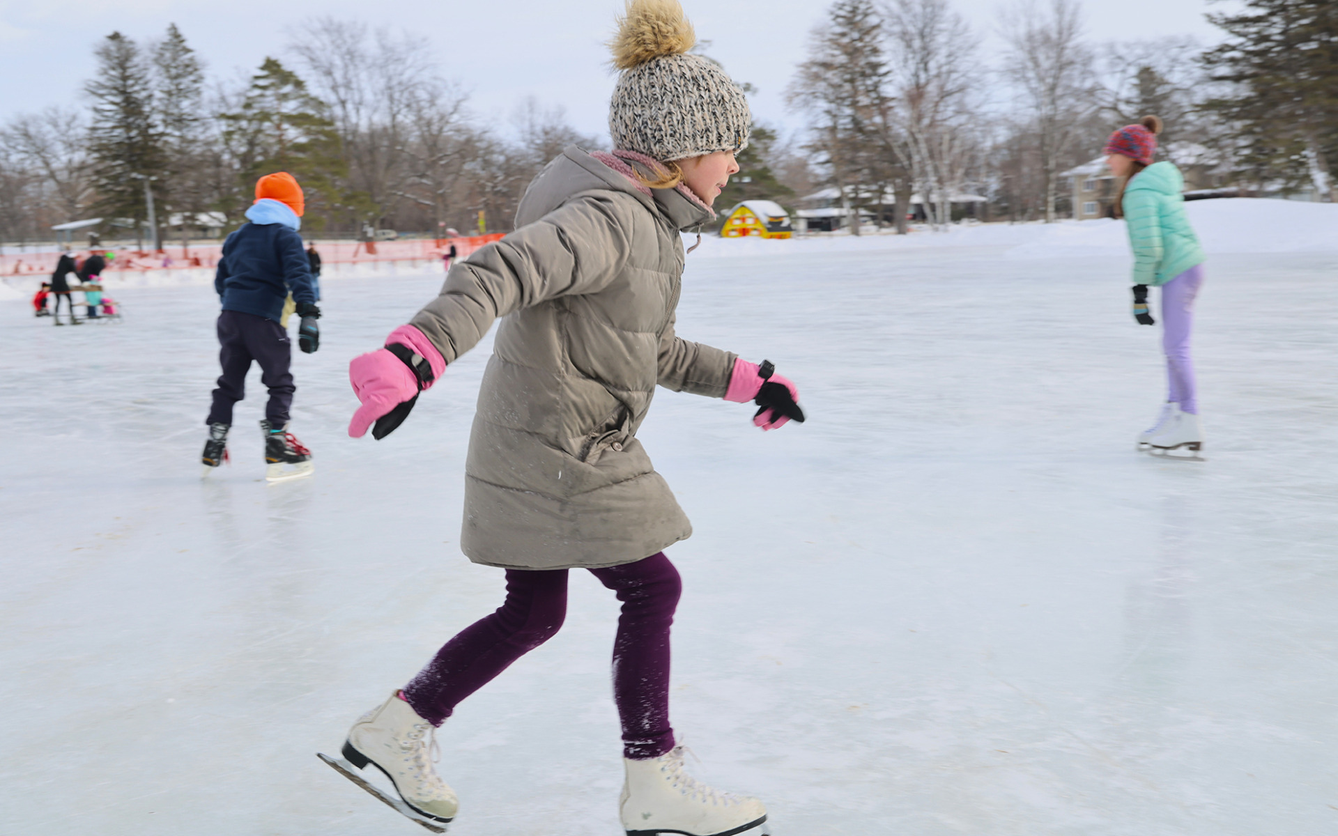 A child in a beige coat, purple leggings, and a knit hat skates on an outdoor ice rink, part of a magical scene from any Winter Guide, with other children in winter clothing gliding across the snowy landscape in the background.