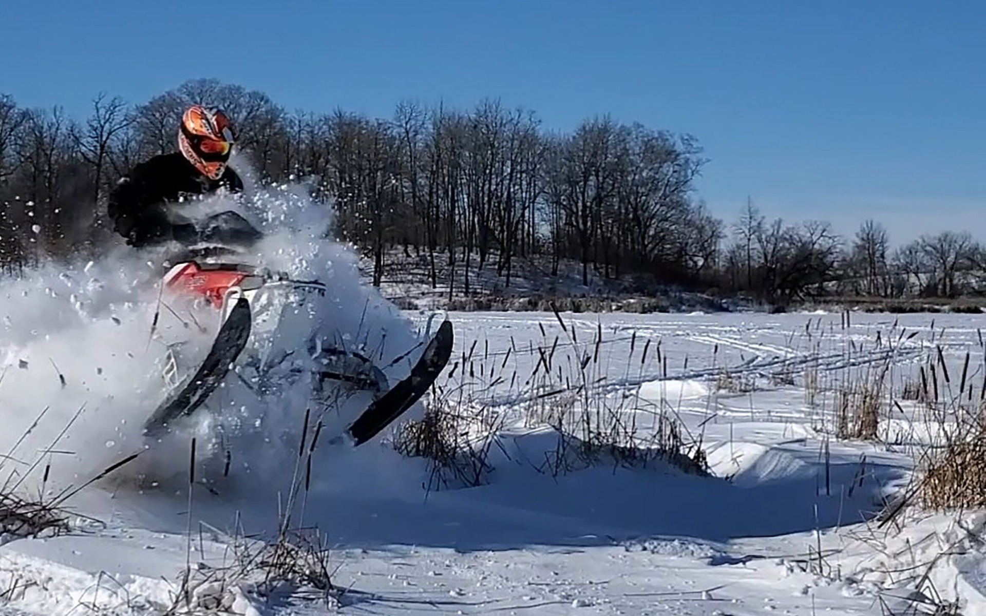 A person in an orange helmet rides a snowmobile at high speed, kicking up snow in the air across a snowy field with leafless trees in the background—a thrilling scene fit for any Winter Guide under a clear blue sky.