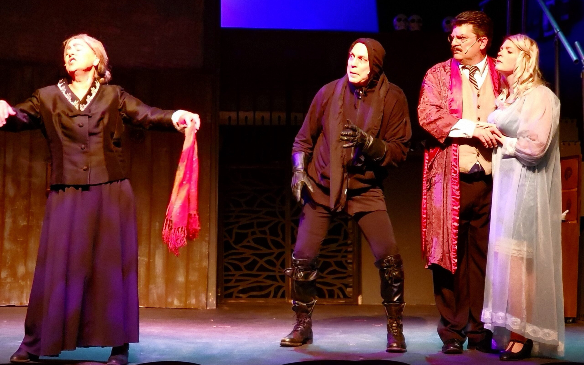 Four actors perform on stage in dramatic costumes; an older woman gestures with a red scarf, a hooded figure crouches in black, and a man in a red robe holds a woman in a light blue nightgown—perfect inspiration for your Winter Guide.