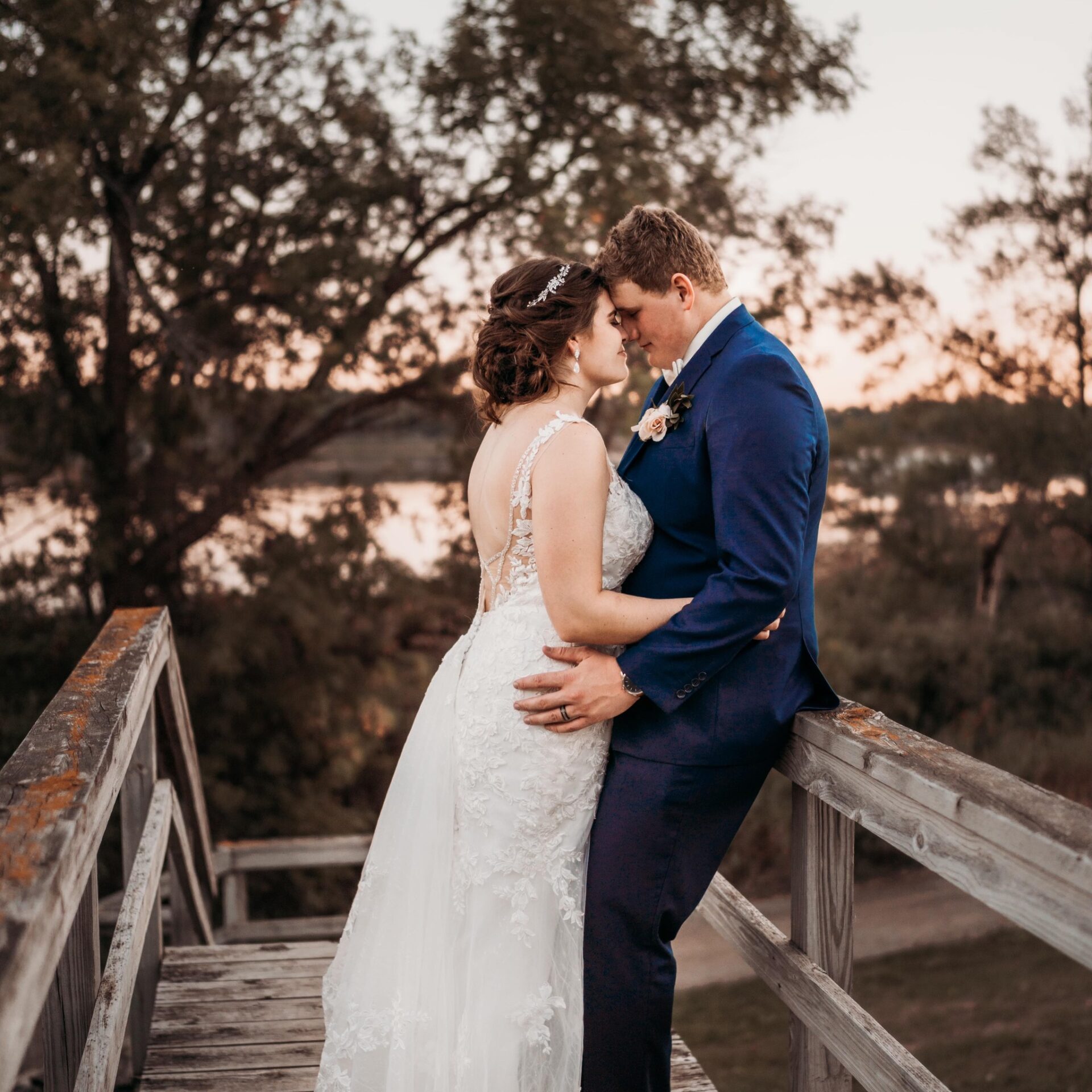 A bride and groom stand close together on a wooden bridge at sunset, touching foreheads and embracing, surrounded by trees with a lake in the background.