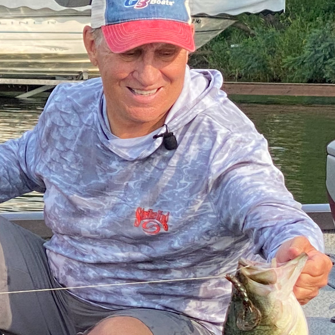 A man in a patterned hoodie and cap, possibly a fishing guide, smiles while holding a large fish on a boat near a dock, with greenery and water in the background.